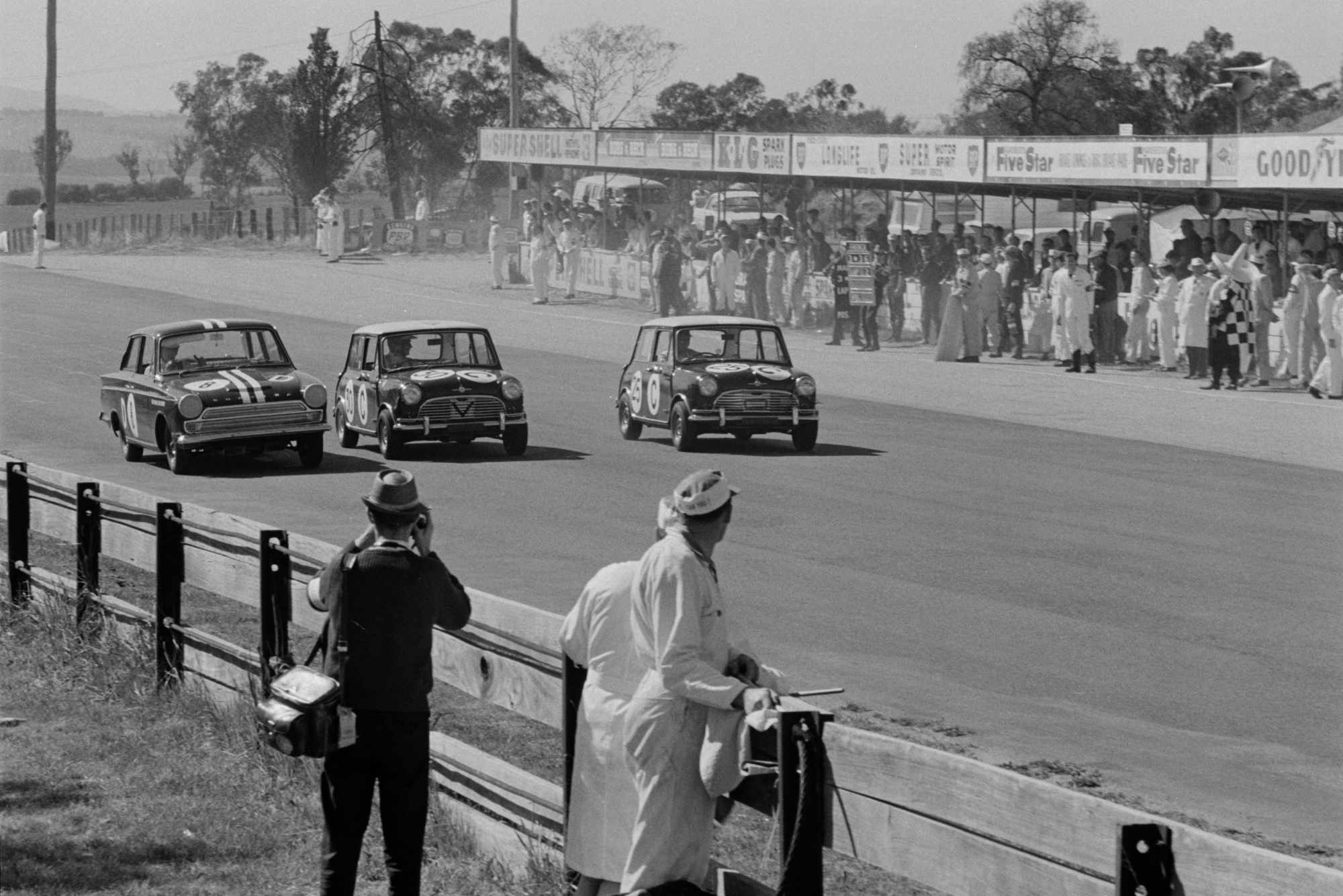 Black-and-white photograph of three racing Minis alongside one another on the start-finish straight at Mount Panorama, Bathurst, during the 1965 Armstrong 500. A larger saloon car leads two Mini Coopers, with spectators, race officials and advertising hoardings lining the pit area. A photographer and officials watch from behind the wooden safety barrier in the foreground.